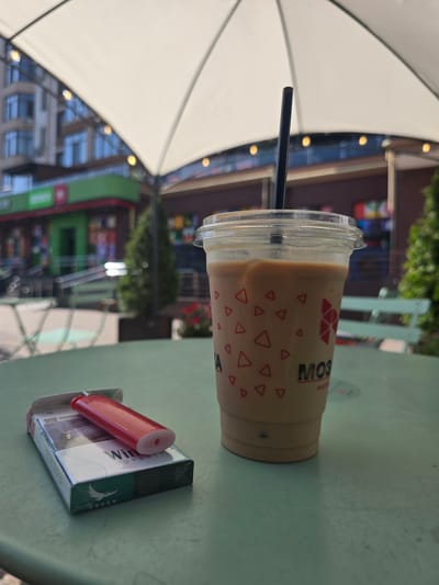 An iced coffee with a black straw sits on a green table next to a pack of "Winston" cigarettes and a red lighter. The table is outdoors, likely on a patio, with a large white awning overhead. In the background, there is a blurred view of buildings and trees, suggesting an urban environment. The context suggests a moment of pause or relaxation. The location is Petropavlivska Borshchahivka, Ukraine.