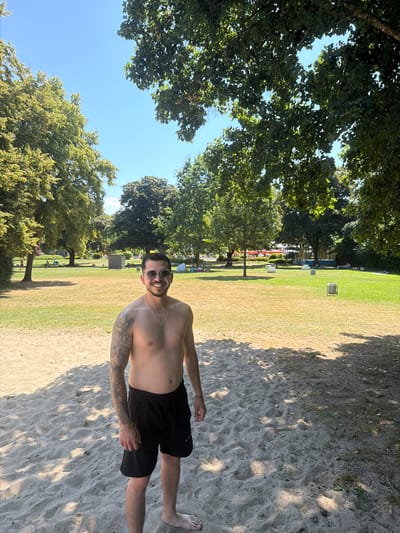 A man with tattoos on his left arm, wearing black swim shorts and sunglasses, stands on a sandy beach. He is smiling and looking at the camera. Behind him, a grassy park area is visible with scattered trees, people relaxing on the grass, and some structures in the distance. The sky is clear and blue. The scene appears to be a public beach or park on a sunny day. This is likely the Emmendingen Stadtpark which features a sandy beach area and grassy spaces for recreation.