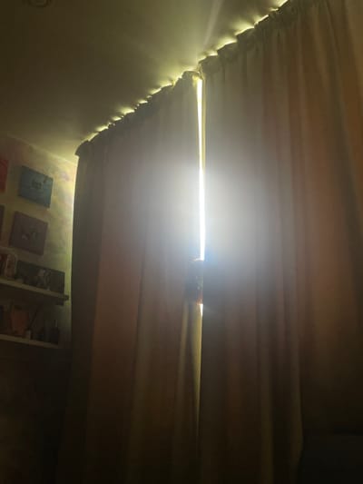 A dimly lit room features closed curtains, with a bright shaft of light shining through a gap in the center. The light is intense and creates a dramatic contrast with the surrounding darkness. On the left wall, several framed pictures are visible, mounted above a shelf. The shelf contains various objects, including decorative bottles and possibly some books. The ceiling is visible at the top of the image and appears to be lit from behind the curtain rod, creating a soft glow. The overall scene suggests a domestic interior setting, possibly in a residential area like ЖК «Борисоглебское», Russia.