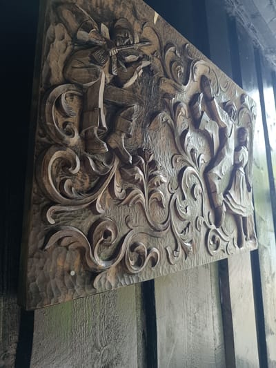 This media file shows a carved wooden panel mounted on a dark wooden exterior wall. The panel depicts a scene with figures and ornate scrollwork. On the left, a figure, possibly a man, is seated and playing a musical instrument that resembles a fiddle or violin. He wears a hat and traditional clothing. To the right of the musician, a second scene unfolds with two figures, a taller figure, likely an adult, standing over a smaller figure, possibly a child. The taller figure appears to be holding the smaller figure, and both are positioned within the elaborate carved floral and scroll patterns that dominate the panel. The carving style is detailed, with a textured background behind the raised figures and motifs. The wood is a warm, natural brown, showing the grain and the marks of the carving tools. The panel is mounted at an angle, suggesting it might be part of a building's facade or a decorative element. The lighting suggests it is daytime, with natural light illuminating the carving and casting shadows. The setting appears to be outdoors, given the visible wooden structure and the natural light. The location context of Hovden, Norway, suggests a rural or traditional setting where such folk art might be displayed. No visible text is present on the carving or its surroundings.
