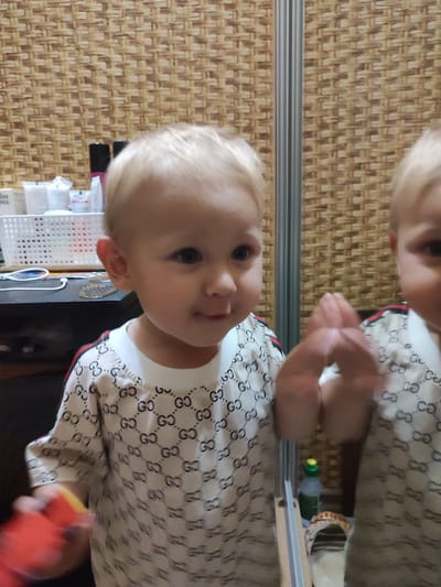 In the foreground, a young child with blonde hair and blue eyes is looking towards the viewer with a slight smile. They are wearing a white t-shirt with a repeating "GG" logo pattern and red and black striped accents on the shoulders. To the right of the child, a reflection of the child and the surrounding environment is visible in a mirror. The background features a woven textured wall and a shelf with various items, including what appear to be bottles and a white basket. A red toy is partially visible in the lower left corner, held by the child. The scene appears to be indoors, likely in a home environment. There is no indication of time of day or weather conditions. The overall mood is casual and domestic. The text "ГЧ" is visible on a bottle in the reflection.