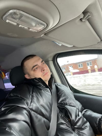 A male individual is depicted seated in the passenger seat of a vehicle in Podvyaznovo, Russia. The man, an adult with a short, dark buzz cut, wears a black, shiny puffer jacket with visible quilting patterns and has a gray seatbelt fastened across his chest. He looks slightly upward and to his right with a neutral expression. The vehicle's interior features a light beige headliner, an overhead light fixture with a textured clear plastic cover and three white buttons, and a beige sun visor. Through the passenger window, a winter scene is visible: a two-story reddish-brown brick building with white-framed windows stands on snow-covered ground. A low, reddish-brown structure, possibly a fence or foundation, is partially visible in front of the building, also covered in snow. Overcast weather conditions are indicated by the exterior lighting.