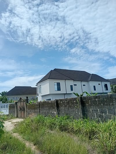 This is a low-angle, medium shot photograph of a residential area in Sangotedo, Nigeria, taken on a cloudy day. The foreground features a rough, dirt path winding through tall, green grass and vegetation. A weathered, concrete block wall runs horizontally across the midground, with a rusty metal gate on the left side of the wall. Behind the wall, two modern, two-story houses are visible. The house on the right is a clean white building with a dark tiled roof and large, dark-framed windows. The house on the left appears older, with a similar dark tiled roof and lighter colored walls, also featuring dark-framed windows. Banana trees and other lush greenery are visible in the background. The sky above is a bright blue, partially obscured by scattered, fluffy white clouds.