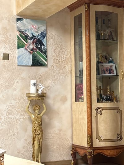 The image shows a wall decorated with a framed photograph of a bride and groom, possibly from a wedding ceremony, placed to the left. To the right, a golden statue of a woman is mounted horizontally on the wall, with her arms extended and her body stretched out. The statue is positioned above a white object with a floral design, which appears to be a candle holder. The wall itself is covered in a light-colored, ornate wallpaper with a repeating damask pattern. Behind the wall-mounted items, a piece of furniture with glass doors is visible, containing various decorative objects, including bottles and framed pictures. The overall scene depicts an interior space, likely a living room or dining area, with a blend of traditional and decorative elements. The lighting suggests it is daytime, with natural light filtering in. The setting appears to be a home environment. No specific weather conditions are discernible. No text is clearly legible in the image, although "MOET" is visible on a bottle within the cabinet. The overall impression is one of a well-decorated and possibly elegant living space.