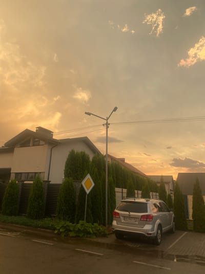 The image shows a residential street in Novi Petrivtsi, Ukraine, at sunset. A silver Dodge Journey SUV is parked in a paved lot on the right side of the image. The car's license plate reads "AA 6663 OX". A row of manicured green trees lines the street, separating it from modern houses. A street light is visible in the center of the image, with power lines stretching across the sky. The sky is filled with dramatic clouds illuminated by the warm hues of the setting sun, casting a golden glow over the scene. A yield sign is present on the left side of the street, indicating a requirement to give way. The road surface appears wet, suggesting recent rain. The overall impression is one of a tranquil suburban evening.