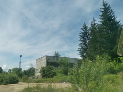 The image depicts an industrial area with an abandoned concrete building and a tall lattice tower in the city of Zhivkovo, Bulgaria. The building, which appears to be a factory or warehouse, has a weathered facade and broken windows. It is surrounded by overgrown vegetation, including trees and bushes. The lattice tower, likely a communication or broadcast tower, stands in the background. The sky is partly cloudy with a few wispy clouds. The foreground shows a dirt or gravel area with some dry grass and weeds. The overall scene suggests a neglected and decaying industrial site within a natural environment.