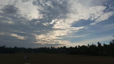 This image depicts a rural landscape under a dramatic sky, likely taken in Buketteukuh, Indonesia. The foreground is a field, dark and appearing fallow or harvested, with hints of some small white markers or flags, one of which is clearly visible. A single dark silhouette of a person can be seen in the distance, and another small, dark figure is near the flag. The middle ground is dominated by a dense line of trees, forming a dark, silhouetted horizon against the sky. Among the trees, some palm trees are identifiable, particularly on the right side of the image. A small, dark building is partially visible within the treeline in the distance.
The sky is the most striking element, filled with a complex arrangement of clouds. Heavy, dark grey clouds are present on the left, obscuring the sun, but allowing shafts of light to break through. Towards the right, the clouds become thinner and more wispy, transitioning to a clear blue sky. The interplay of light and shadow suggests either dawn or dusk. The overall mood is serene and perhaps a little melancholic due to the heavy clouds and dark landscape, but also hopeful with the breaking sunlight. There is no visible text in the image.