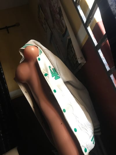 The scene is an interior location in Oba-Ile, Nigeria. A light brown, headless, armless female mannequin torso is positioned diagonally, ascending from the lower left to the upper right of the frame. A white varsity jacket, featuring green trim, green snap buttons, and green embroidered lettering (likely 'LA') on the left chest, is draped open over the mannequin torso. The upper left background shows a yellow-tan wall section with a dark horizontal rod. The upper right background displays a window secured by dark metal bars, bordering a reddish-brown wall section. Natural light enters primarily from this window. Behind the mannequin, a vertical white fabric banner with dark text, including "CHEF" and "MILK," is visible on the wall, alongside a partially obscured, faded poster. The overall illumination is dim. No individuals are present.