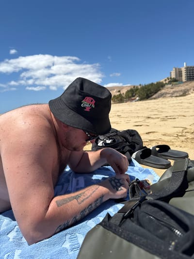 This image, currently displayed on its side, depicts a person relaxing on a sandy beach under a bright, clear blue sky in Pájara, Spain. The central figure is a shirtless man, seen from the chest up, lying on a light blue towel. He is wearing black sunglasses and a black bucket hat. The hat features a pink logo with text that appears to read "DUCK SKATE CONTEST."
His arms are visible, displaying several tattoos, including a knuckle duster design on his left hand and a stylized knife or dagger on his left forearm. Around him, various personal items are scattered on the towel and sand: a pair of black slides, a black duffel bag or backpack, and a pack of "Smoking Deluxe" rolling papers.
The background reveals a wide expanse of light brown sand, leading to distant greenery and a tall building, possibly a hotel or resort, under a sky dotted with a few white clouds. The bright sunshine indicates it is daytime, suggesting good weather for leisure and relaxation on the beach.
