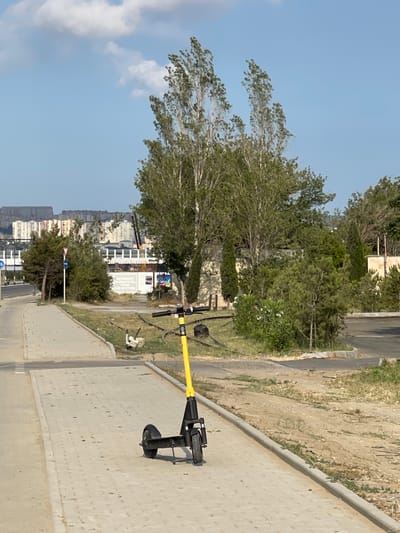 The image depicts an outdoor scene on a bright, sunny day. A yellow electric scooter stands upright on a paved walkway. The scooter is black with a yellow handlebar and has a wheel visible at the back.
Beyond the walkway, there is a strip of dry, grassy terrain next to a road. Further in the background, lush green trees and vegetation are abundant. A low-rise building with white walls and several windows is visible behind the trees. In the far distance, a cityscape with numerous tall apartment buildings can be seen under a clear blue sky with scattered clouds.
The time of day appears to be daytime, likely late morning or afternoon, given the bright sunlight and clear sky. The weather is fair and sunny. The presence of the electric scooter suggests an urban or suburban environment where such transportation is common. The mixture of natural greenery and urban development indicates a developed area. No people are visible in the frame. The scene is calm and static, with no apparent activity taking place.