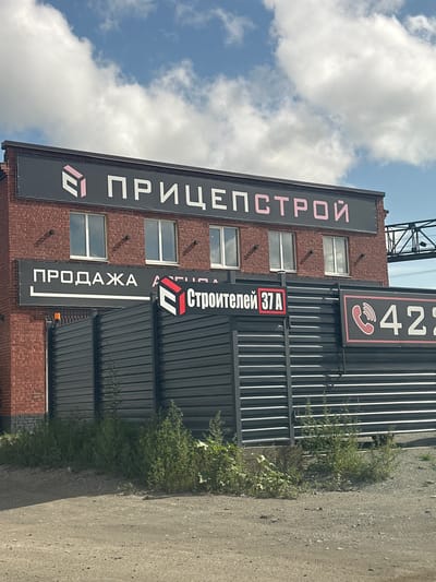 The image shows the facade of a brick building with signage. At the top, a black sign reads "ПРИЦЕПСТРОЙ" in pink letters, with a pink and white geometric logo to its left. Below this, a smaller black sign says "ПРОДАЖА" (sales) in white letters. To the right of "ПРОДАЖА", partially obscured by a fence, is another sign with a geometric logo and "Строителей 37А" (Builders 37A). A black corrugated metal fence runs along the front of the property, and to the right of this, another black sign features a red telephone icon and the numbers "422". The building has multiple windows, and the sky above is partly cloudy. The foreground shows a dirt and gravel surface. This scene is likely in Orsk, Russia, given the Russian text on the signs.