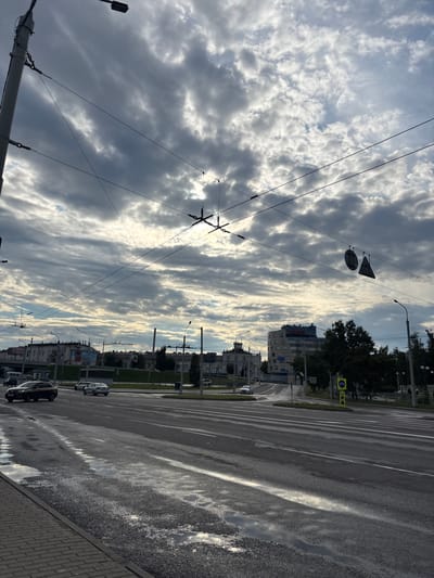 The image depicts a street scene in the vicinity of Сеніцкі сельскі Савет, Belarus. The sky is heavily clouded with streaks of sunlight breaking through. Overhead, a network of trolleybus wires crisscrosses the frame, suspended from poles. In the foreground, a wet asphalt road reflects the sky and surrounding light. Several cars are visible on the road, including a yellow taxi. The background features buildings, some with modern architecture and others appearing older, alongside trees and streetlights. Traffic signs, including pedestrian crossing and directional signs, are present. Puddles on the road suggest recent rain.