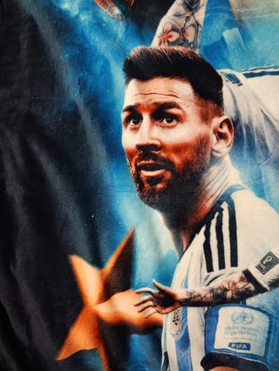 This image, taken in Sucha Beskidzka, Poland, features a close-up view of a graphic printed on fabric, likely a blanket or poster. The graphic depicts a stylized portrait of a man, who strongly resembles Lionel Messi, a famous soccer player. He is shown looking to the right with his mouth slightly open. He is wearing a blue and white striped shirt, characteristic of the Argentine national team, and has a beard and tattoos on his arm. The background is a dark blue gradient with scattered light specks, evoking a cosmic or celestial theme. A bright, orange, star-like shape is positioned to the left of the man's torso, partially obscuring his lower body. A blue band with the "FIFA" logo and "World Health Organization" text is visible on his upper arm. The lighting is dramatic, with the man's face illuminated from the front, highlighting his features and casting subtle shadows. The texture of the fabric is visible, suggesting a soft, plush material.