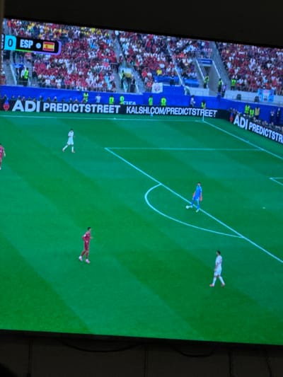 The image captures a screen displaying a soccer match. The scoreboard at the top left indicates "0 ESP," suggesting a score of zero for Spain. A small Spanish flag is visible next to the score. The field is green with white markings, and several players in white and red uniforms are present. A goalkeeper in a blue uniform is near the penalty box. The background shows a stadium filled with spectators. Along the bottom of the screen, a banner displays text including "ADI PREDICTSTREET" and "KALSHI.COM/PREDICTSTREET." The scene is likely at a sports stadium in El Escorial, Spain, given the indication of "ESP" for Spain on the scoreboard.