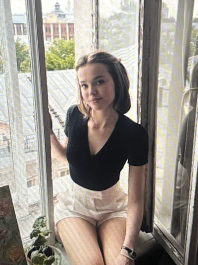 The image shows a young woman sitting on a windowsill, facing the camera. She has brown hair, pulled back in a half-up style, and is wearing a black v-neck top and white shorts. Her left arm is resting on the windowsill, and her right hand is visible on her lap. In the background, through a window, are buildings and trees, suggesting an urban setting. The woman is positioned in the center of the frame, with a window on either side of her. There is a potted plant on the windowsill to her left.

Based on the provided information, the location is identified as Pruszcz Gdański, Poland. This detail is not visually evident from the image itself, but is attributed to external context.