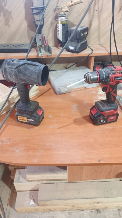 The image depicts a workbench cluttered with tools and materials. In the foreground, two cordless drills are prominent: one black and grey on the left, and a red and black one on the right. The drill on the left has a wide, funnel-like attachment. The drill on the right appears to be actively drilling, with a white rod protruding from its chuck. Behind the drills, on a shelf, are various items including a can of spray, what appears to be a soldering torch, and battery packs. The workbench is made of wood and shows signs of use and dust. Below the workbench, stacked pieces of lumber are visible. The overall environment suggests a workshop or garage setting. The image is focused on the tools and the immediate workspace. No specific location in Pavino, Russia can be identified from the visual evidence presented.