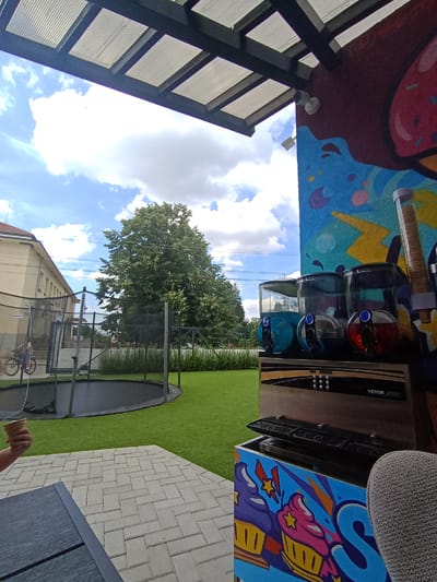 In Konstantinovo, Bulgaria, an outdoor area features a trampoline, a building, and a slushy machine. The scene is captured under a partly cloudy sky. A person's arm is visible, holding an empty ice cream cone. The slushy machine, a VEVOR model, has three dispensers, one filled with blue liquid, and another with red. The machine is decorated with colorful cartoon cupcakes. A graffiti-style mural adorns the adjacent wall, depicting ice cream cones and abstract designs. The ground is paved with light-colored interlocking bricks. A dark-colored table is partially visible in the foreground.