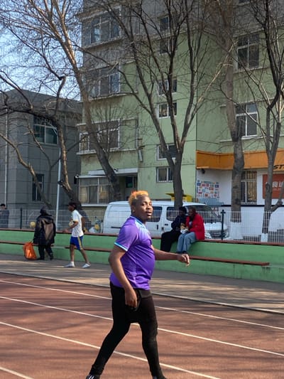 The image displays an outdoor scene within Taihe District, China, under a clear sky. In the foreground, a running track with reddish-brown surfacing and white lane lines is visible. A male subject with bleached blonde hair, wearing a purple and light blue athletic shirt and black pants, walks from left to right across the track, his head turned slightly to the left. In the mid-ground, another individual in a white shirt and blue shorts walks right on the track. Further left, a person in a dark coat walks away from the camera, carrying an orange bag near a yellow ball. Behind the track, a low green and brown bench or wall separates the track area from a paved section. Two individuals are seated on this bench: one in dark clothing, and another in a red jacket and blue jeans, holding a food item. A white panel van is parked behind the seated individuals. The background features several multi-story buildings, including a grey structure on the left and a light-green building on the right with an orange awning and various commercial decals on its ground floor. Bare-branched deciduous trees are interspersed across the scene, partially obscuring the buildings.