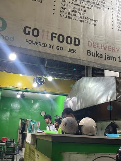 This is a photograph taken at an outdoor food stall or small restaurant, likely in South Tangerang, Indonesia. The scene is dimly lit with artificial lights, suggesting it could be evening or night. The main focus of the image is a large banner advertising food items and delivery services. The banner is mostly in Indonesian and features the "GO FOOD" logo, indicating it's a partner with a food delivery platform. Visible text on the banner includes food names like "Nasi Bistik Ayam," "Nasi Bistik Sapi Mentega," and "Nasi Bistik Sapi Tepung," as well as operational hours: "Buka jam 1" (Open at 1 o'clock).
In the background, several people are visible. Two young men appear to be working behind a counter, with one looking down at papers. The setting includes a green wall, and various objects like motorcycle helmets (one white, one grey) are displayed on a shelf. The overall atmosphere seems functional and typical of a local food establishment.