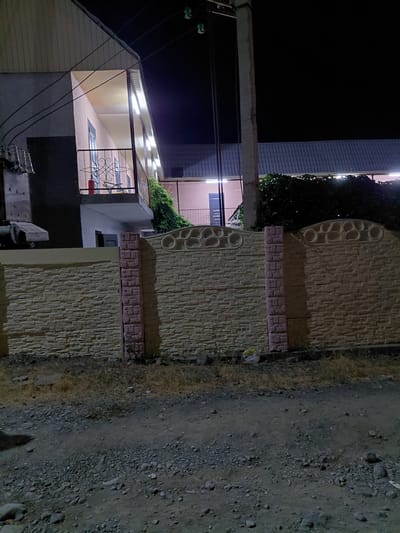 The image shows a nighttime view of a building and a decorative stone fence in Ақши, Kazakhstan. The building appears to be a house or a small business, with a roofline illuminated by bright white strip lights. A covered porch or patio area is visible, with a metal railing and a glimpse of red and blue objects inside. The fence is made of light-colored textured stone with decorative circular patterns and a band of smaller, darker square bricks. To the right of the fence is a rough, gravelly ground surface. The sky is dark, indicating it is nighttime. There are no people visible in the image. The scene appears quiet and still. The overall impression is of a residential or commercial property at night.