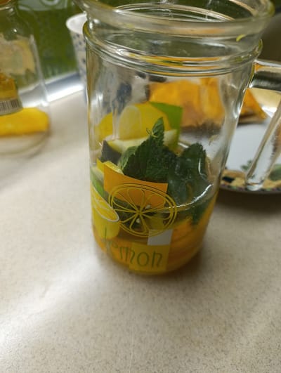 A close-up shot captures a glass pitcher filled with a beverage infused with citrus slices and mint leaves. The pitcher has a decal of a lemon slice on its exterior. Behind the pitcher, a partially visible bottle contains a green liquid, and a white cup with a blue pattern is also present. The scene appears to be indoors, set on a speckled countertop. The lighting is somewhat dim, creating reflections on the glass. There is no discernible connection to the city of Pivdenne, Ukraine, in this image.