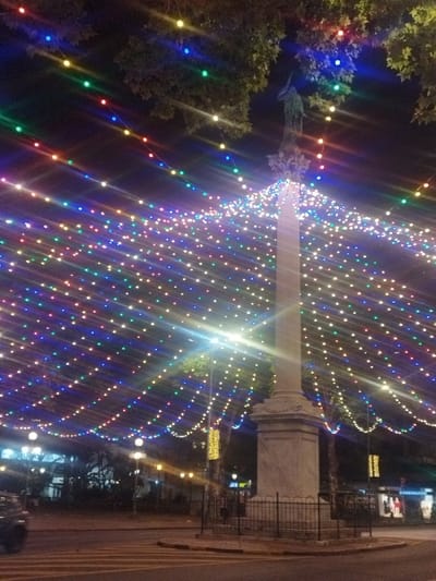 A brightly illuminated night scene features a tall stone monument with a statue atop, surrounded by numerous strands of colorful lights strung overhead. The lights form a canopy effect, creating a vibrant and festive atmosphere. Trees with dark green foliage frame the upper portion of the image, adding depth. Below, a city street with building facades and street lamps can be seen in the background, hinting at an urban setting.