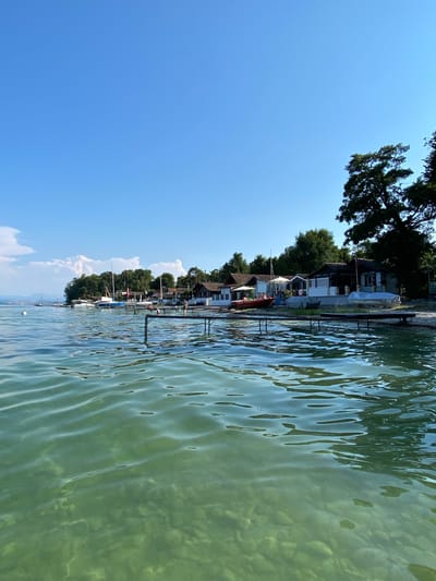This image depicts a scenic view of a waterfront in Milvignes, Switzerland, on a clear day. The foreground is dominated by the tranquil, turquoise waters of a lake, revealing a rocky bottom due to its clarity. Gentle ripples on the water's surface reflect the bright sunlight.
Along the shoreline, a row of small, white and brown houses with peaked roofs are visible, suggesting a residential or recreational area. In front of the houses, wooden piers extend into the water. Several boats, including sailboats and smaller motorboats, are moored near the shore and along the piers. Some boats are covered with tarpaulins.
A diving platform with metal supports is also present, and two figures are seen on the platform, with one appearing to be about to jump into the water, and the other already in the water. Lush green trees line the background, providing a verdant backdrop to the structures and the water. The sky is a clear, vibrant blue, with a few wispy clouds scattered in the distance. The overall atmosphere is one of peaceful summer leisure.