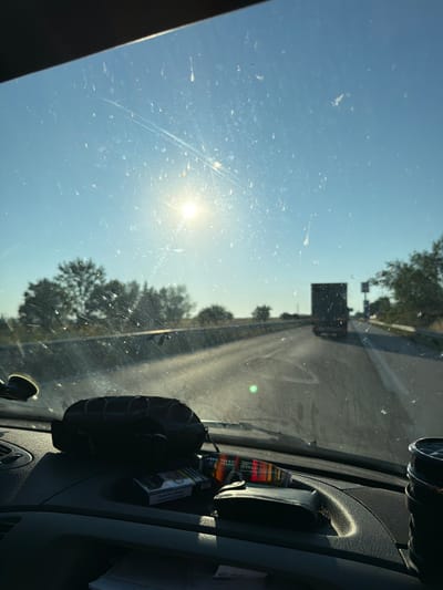The image depicts a view from inside a vehicle, looking through the windshield. The windshield is dirty and smudged, with streaks and spots visible. The sun is shining brightly in the sky, causing lens flare and obscuring details. In the distance, a dark truck is driving on a highway. Trees line the sides of the road.
In the foreground, on the dashboard, are several items. A black pouch with a patterned drawstring is positioned towards the left. Next to it is a pack of cigarettes. To the right of the cigarettes is a wallet or small pouch with a striped patterned edge. A black cup holder or container is visible on the far right. The dashboard itself appears to be made of dark, textured plastic.
The location is specified as Orizovo, Bulgaria. The scene is consistent with a highway drive in a rural or suburban area, given the presence of trees and a truck on the road. The time of day is likely morning or afternoon due to the bright sun and clear sky. No people are clearly visible within the vehicle.