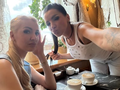 Two women are taking a selfie at a cafe table in Novaya Usman', Russia. The woman on the left, with blonde hair in a ponytail, wears a light blue top and a watch. The woman on the right, with dark hair, wears a white tank top with rhinestones and a necklace. She is leaning towards the camera and puckering her lips. In front of them are two cups of coffee on a table with a marble-like pattern. Behind them, a window with sheer curtains reveals greenery and a cloudy sky. A wall sconce with crystals is visible on the right wall.