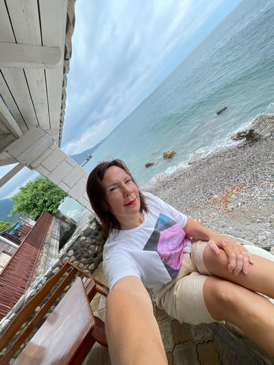 A woman is taking a selfie outdoors, sitting on a bench. She is wearing a white t-shirt with a pink and black graphic and beige shorts. She has brown hair and is wearing red lipstick. The background shows the sea and a pebble beach. To the left of the frame is a wooden structure with a white roof and a stone wall. Further to the left, there is a red-roofed building and a green tree. In the distance, mountains and the coastline are visible. The location is Abaata, Abkhazia.