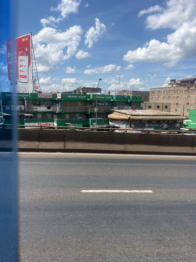 The image depicts an urban street scene in Githurai, Kenya, under a partially cloudy blue sky. In the foreground, a multi-lane asphalt road, partially obscured by a blue vertical blur on the left, features a single white dashed line. This road is separated from the background by a horizontal concrete barrier.
Beyond the barrier, the midground is dominated by a row of multi-story buildings. The most prominent are residential structures painted predominantly green, featuring multiple balconies with visible laundry hanging. These buildings display prominent "Safaricom m_pesa" and "Bata" signage. To the right of these green buildings, a lower, single-story structure with a corrugated metal roof and multiple covered window openings features murals or graffiti on its façade. To the far left, a large billboard stands tall, advertising "Colgate Brush and unlock Rewards" and "Naivas." Streetlights are visible above the buildings. The background reveals additional multi-story buildings, appearing older and constructed from brick or unpainted concrete. No individuals or explicit actions are clearly discernible in the scene.