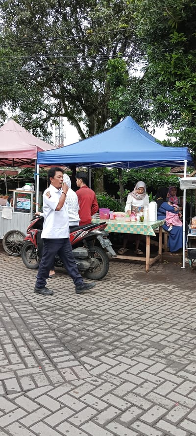 The image shows a street scene in Banjaran, Indonesia (latitude: -7.046, longitude: 107.581). A man in a white shirt and black pants walks past a motorcycle parked in front of a blue canopy tent. The man is holding a phone to his ear. Inside the tent, there is a table with food and drinks, and a woman in a headscarf is serving customers. Two other women are seated at the table, and a man is standing behind them. The street is paved with concrete blocks. The scene is under a partially overcast sky. Several power lines run across the top of the image. There is a pink canopy tent to the left of the image.