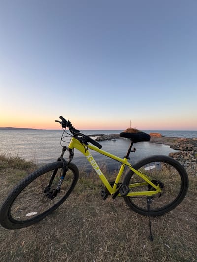 A bright yellow mountain bike is parked on a grassy embankment overlooking a calm body of water at sunset. The sky transitions from a pale blue at the top to soft oranges and pinks near the horizon. The water stretches out to meet the distant shoreline, which is marked by a rocky outcropping. On this outcropping, a small harbor with a few boats is visible, along with a prominent cross on a small hill. The bike is positioned in the foreground, with its front wheel angled towards the left and its back wheel towards the right, resting on its kickstand. The bike's frame displays the "X-RULER" logo. The surrounding ground is covered in dry, straw-colored grass. The overall scene suggests a peaceful moment in a coastal location, likely Lozenets, Bulgaria, given the presence of a cross on a hill which is a common landmark in the region.
