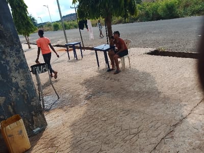 The image depicts a street scene in Umueshi, Nigeria. Two young people are visible in the image. One person, wearing an orange shirt, is carrying what appears to be a portable charcoal grill. The other person is sitting on a white plastic chair at a blue table, talking on a cellphone. In the background, laundry is hanging on a line. The scene appears to be on a paved area alongside a road.