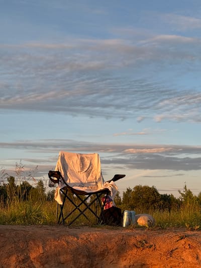 An empty camping chair draped with a white sheet sits in a field near Sapunov Bor, Russia, at dusk. The chair is positioned on a dirt embankment, with tall grass and trees in the background. A bag, two cans, and a light-colored object, possibly a rolled-up blanket or sleeping bag, are placed on the ground near the chair. The sky is a pale blue with streaks of pink and gray clouds. The lighting suggests the setting sun, casting a warm glow on the scene.