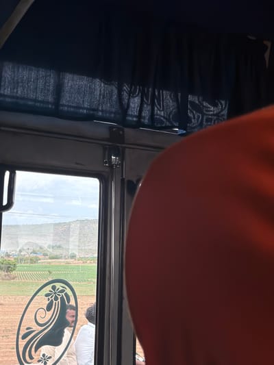The image shows the interior of a vehicle, possibly a bus, looking out a window. A person's arm, with skin illuminated by a reddish hue, is visible in the upper part of the frame, suggesting the photographer's presence. To the left, dark blue curtains with a textured pattern are drawn, allowing some light to filter through.
The main focus of the image is the window, which offers a view of an outdoor scene. The landscape outside appears to be rural, with fields of crops in various shades of green and brown, stretching towards a distant, hazy mountain. The sky is overcast, with diffused light suggesting it might be daytime, though the exact time is unclear. The weather appears to be fair, with no visible rain.
On the glass of the window, there is a dark, decorative decal featuring stylized floral and swirling patterns, possibly a silhouette of a woman's profile. Reflected in the window, we can see a glimpse of two people. One person is wearing a white shirt and appears to be leaning forward. The other person's face is partially visible, and they seem to be looking out the window as well. The setting is likely Thammarajupalle, India, based on the provided location context. The overall mood of the image is calm and observational, capturing a moment of travel and the scenery outside. No visible text is present in the image.