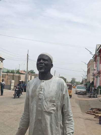 An outdoor scene in Hotoro South, Nigeria, depicts a dark-skinned adult male in the foreground, positioned slightly right of center, looking upwards and to his left. He wears a light-colored, vertically striped tunic with a chest pocket and four visible buttons, along with a light-colored skullcap. He has a short, patchy grey beard. The subject stands on an unpaved road.
In the midground, a two-person motorcycle travels away from the viewer on the left side of the road. Further left, a male in a white shirt walks. A building under construction with exposed concrete blocks and a green-roofed structure are visible beyond. Utility poles with overhead wires cross the upper portion of the frame.
The right side of the road extends into the distance, lined with buildings featuring red accents and external air conditioning units. Several individuals are visible sitting or standing along the street. A pile of large root vegetables is present on the ground near the right edge. Vehicles on the road include a yellow auto-rickshaw and a silver sedan. Streetlights are mounted on poles along the thoroughfare. The sky is overcast or hazy.
