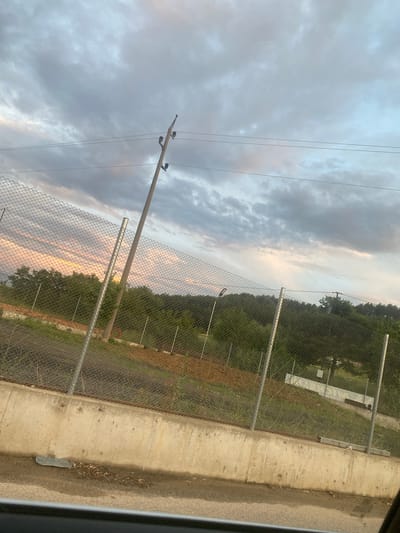 The image shows a cloudy sky with a sunset casting pink and yellow hues over the horizon. In the foreground, a chain-link fence runs horizontally across the frame, supported by metal posts. Behind the fence, there is a concrete retaining wall, and beyond that, a gently sloping hill covered in green trees. A wooden utility pole with insulators and wires stands diagonally against the sky. The scene appears to be on the outskirts of Ablanitsa, Bulgaria, possibly near a road given the view from a vehicle. There are no people or animals visible in the image. The overall impression is one of a rural or semi-rural landscape at dusk.