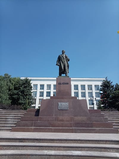 The image shows a statue of Vladimir Lenin in the city of Zheleznovodsk, Russia. The statue stands on a large granite pedestal in front of a white building with many windows. Trees surround the monument. The sky is clear and blue. The photo is taken from a low angle, looking up at the statue.