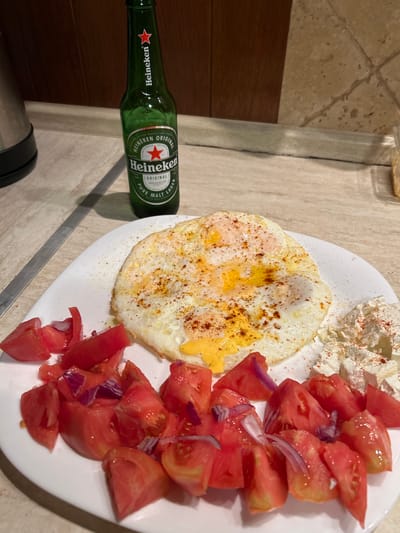 This is a close-up, eye-level, indoor photograph of a meal on a white plate, accompanied by a bottle of Heineken beer. The meal consists of two fried eggs with runny yolks, seasoned with paprika and pepper. To the left of the eggs are chopped tomatoes mixed with red onion. To the right of the eggs is a pile of crumbled white cheese, also sprinkled with paprika. The Heineken bottle, a standard green glass bottle with its label clearly visible, is positioned behind the plate. The setting appears to be a kitchen counter, with a tiled backsplash in the background and a wooden cabinet to the left. A metal kettle is partially visible on the far left. The lighting is natural and slightly warm. The scene does not depict any people, specific actions, or interactions. The location is not identifiable as Polenitsa, Bulgaria.