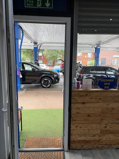 The image shows a view from an open doorway, looking out onto an outdoor car wash facility in Salford, United Kingdom. In the foreground, a brown coir doormat is visible on a grey wooden floor. Beyond the doormat, there is a patch of artificial green grass.
The car wash area features a silver-grey concrete floor with yellow markings. Several cars are present, including a black SUV with distinctive wheels on the left, and a black hatchback with a large spoiler on the right. A person wearing a black cap and a grey vest is standing near the black hatchback, appearing to be interacting with the car. Another person, dressed in dark clothing, is visible standing by the black SUV.
Overhead, a white corrugated roof structure is supported by blue posts. Various hoses and cables are suspended from the roof. Signs are visible in the background, including a green "Fire exit" sign with a running man and arrow symbol, and a blue oval sign on the right that reads "SCREEN WASH TOP UP HERE". Two plastic bottles, one clear and one pink, are placed on a wooden counter in the foreground of the car wash area. The buildings in the background appear to be brick residential properties.