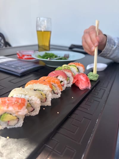 A close-up, eye-level shot captures a dining scene featuring a variety of sushi rolls on a dark, rectangular platter. The image is taken in what appears to be a restaurant or cafe in Hainburg an der Donau, Austria.
In the foreground, a person's hand, partially visible and wearing a light-colored denim jacket, holds a pair of wooden chopsticks. The chopsticks are poised over a small dollop of green wasabi, indicating the person is about to add it to their meal. Adjacent to the wasabi is a vibrant pile of pink pickled ginger (gari) and a white ceramic chopstick rest.
The sushi rolls are neatly arranged in a long line, showcasing different types, including maki rolls topped with fresh salmon, tuna, shrimp, and avocado, as well as rolls containing crab stick and sprinkled with sesame seeds. The dark platter has an embossed geometric pattern, possibly a Greek key design, running along its edge.
In the blurred background, a tall glass filled with a golden liquid, likely beer, is visible. To the left of the sushi plate, a light blue-grey plate holds a small portion of green salad and a fork. A red can with indistinct white text (possibly "Amstel" or a similar brand, though not clearly legible) and a dark menu or booklet are also present on the table. The overall impression is a casual yet appealing meal.
