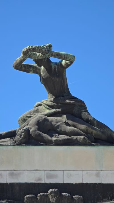 The image presents a close-up, low-angle view of a monumental bronze statue, displayed here rotated 90 degrees counter-clockwise from its upright position. The statue, which depicts a classical human figure (likely female) with a slender form, stands with both arms raised above its head, holding a substantial laurel wreath. The figure is draped in flowing robes, revealing sculpted contours and details. The bronze material shows a rich, weathered greenish-blue patina, indicative of age and exposure.
This sculpture is an integral part of a larger monument, with a smooth, light-colored stone or marble section visible to the left (which would be the base if the image were correctly oriented) and other dark, textured sculptural elements to the right. The entire scene is set against a clear, brilliant blue sky, suggesting bright daylight. A faint airplane contrail can be observed in the sky. The strong sunlight creates distinct shadows, highlighting the statue's three-dimensional details. No text is visible on the monument.