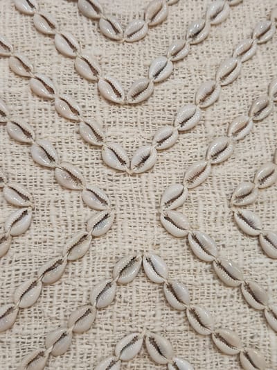A close-up, overhead view of a natural fiber textile with a decorative pattern of small, white cowrie shells strung together on thin white thread. The shells are arranged in diagonal lines forming a diamond-like pattern across the fabric. The textile appears to be a loosely woven, textured material, possibly linen or a similar natural fiber, in a light beige or off-white color. The shells themselves are smooth and glossy, with a characteristic dark opening. There are no people or other objects visible in the image. The lighting is even, casting minimal shadows, highlighting the texture of both the shells and the fabric. The image is sharp and in focus, allowing for clear detail of the individual shells and their arrangement. The location in Maitland, United States of America, is not evident from the visual information present in the image.