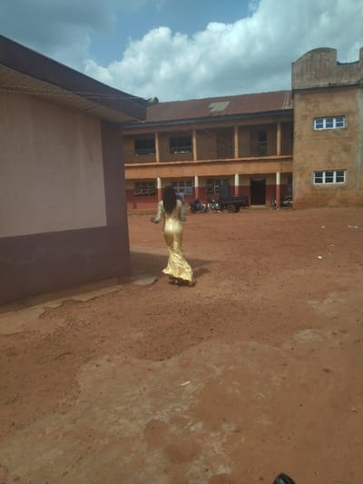 A woman in a long gold dress walks away from the camera in a dirt courtyard. The courtyard is between a two-story building with a brown roof and a one-story building. The ground is dirt. There are two motorcycles parked on the left side of the image. The coordinates (latitude: 7.503, longitude: 7.174) place this image in Anyigba, Nigeria.