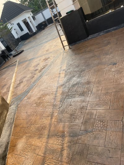 The image displays an outdoor paved area in Ogbunu, Nigeria. The ground is covered with stamped concrete featuring tan and brown tones, patterned to resemble a combination of wood grain and pebble textures arranged in interlocking rectangular blocks. Dark gray concrete strips form geometric divisions across the pavement. To the left, a white single-story building with a dark gable roof, black window frames, and a black lower facade is visible, with another similar structure partially seen behind it. A dark chair and a plant in a dark circular planter are positioned near the foreground building on the left. On the right, a multi-rung metal ladder leans against a white structure with a black lower section, casting a prominent shadow across the patterned concrete. A small, light-colored triangular frame rests on the pavement in the lower-left corner. The scene is brightly illuminated by natural light, evidenced by distinct shadows. No individuals are present in the image.
