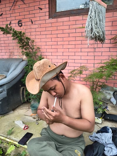 In An Khe Ward, Vietnam, a shirtless male, wearing a brown cowboy hat and green sweatpants, is seated cross-legged on a concrete surface. He is actively lighting a white-tipped cigarette held in his right hand using a lighter in his left hand, with his gaze directed towards the cigarette. A black smartphone and a pair of white work gloves are positioned on the concrete to his left. Behind him, a dark, worn sofa fragment leans against an unseen object, with a small green bucket partially visible. The background consists of a reddish-orange brick wall featuring large, dark numerals (including '6' and '1') and markings suggestive of an outdoor clock, and a wooden-framed window. A used string mop hangs from the wall to the right of the window. Various green plants are present in the foreground and background. A pile of dark clothing is located on the ground to the individual's immediate right.