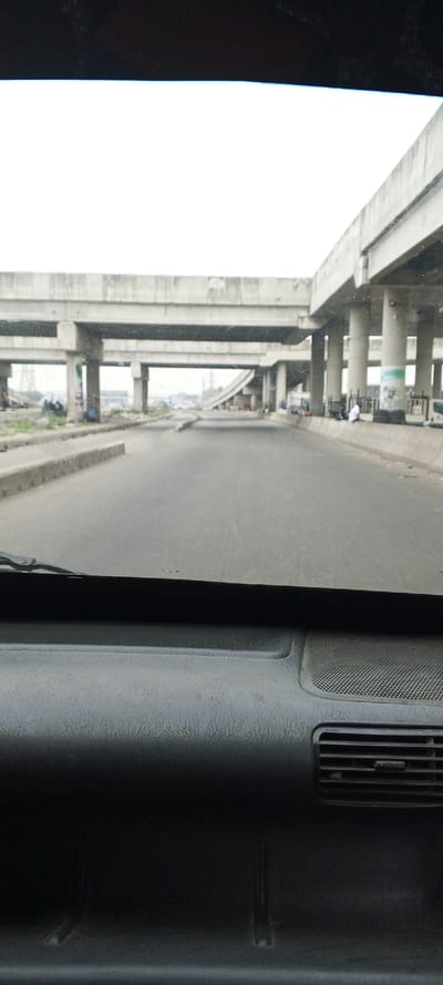 The image depicts an urban road scene, viewed from the interior of a vehicle, within the city of Road, Nigeria. The foreground shows the vehicle's dark, textured dashboard with a visible air vent on the right, and a partial view of a windshield wiper blade on the upper left. A grey asphalt road occupies the midground, stretching forward under an extensive network of concrete flyovers and expressways. A low concrete barrier separates the road on the left.
Multiple concrete overhead bridges, supported by thick pillars, span the roadway. On the right side, another elevated concrete structure curves away, with a visible green and white poster or sign attached to one of its support pillars. Beneath these elevated structures, on both sides of the main road, human presence and activity are evident. On the right, several individuals are observed, with one person wearing a light-colored garment seated or crouching near a concrete pillar. On the left, indistinct figures and what appear to be informal structures or gathered materials are present. The sky is uniformly bright and overcast.