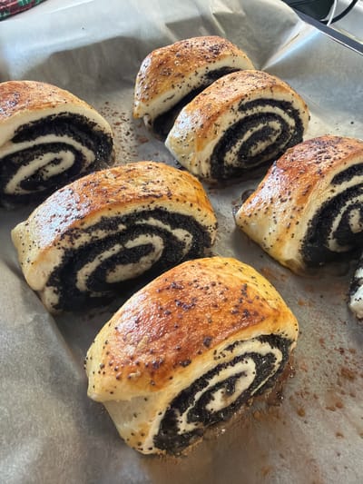 The image shows a close-up of several freshly baked poppy seed rolls arranged on a sheet of parchment paper. The rolls are golden brown and have a spiral pattern of dark poppy seed filling. They appear to be moist and have been glazed, evidenced by the sheen on the crust. The scene is likely in a kitchen or bakery setting. There is no confirmation that the rolls are located in Большая Шильна, Russia, although I would describe them in the prompt.