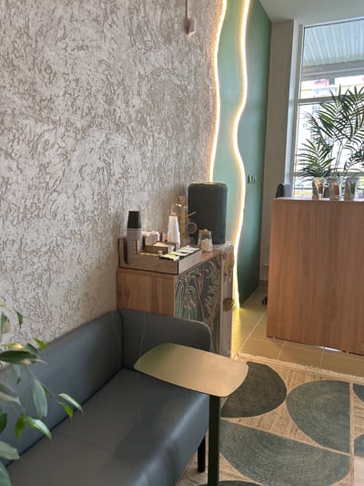 The image depicts an interior scene, likely a cafe or waiting area, in Medvedevo, Russia. The left side of the frame shows a textured, off-white wall with a decorative, wavy vertical light installation emitting a warm glow. In the foreground, a dark gray sofa with visible stitching detail is partially visible, with a small, metallic side table positioned on its right.
To the right of the sofa and wall, a wooden cabinet stands. On top of this cabinet is a wooden organizer holding stacks of paper cups and what appear to be sugar packets. A water cooler with a gold dispenser is positioned to the left of the cabinet. Behind the cabinet, a large, dark green object, possibly a cushion or upholstered item, is placed against the wall.
Further to the right, a wooden counter or desk is visible, with several clear glasses containing palm fronds. A large window is present on the far right, showing a view of buildings and foliage outside. The floor is tiled with light brown square tiles, and a rug with a geometric pattern of large, overlapping oval shapes in shades of beige and dark green is laid out in the foreground. The overall impression is of a modern, clean, and well-lit space.
